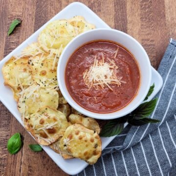 A white plate with air fried ravioli and a bowl of marinara sauce