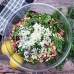 An overhead photo of Mediterranean Tomato Cucumber Salad in a clear bowl, accessorized with a black and white napkin, two lemons, and dill and parsley sprigs