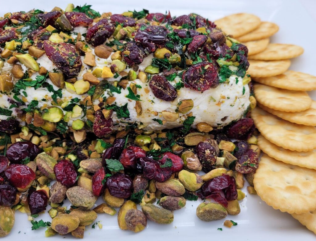 A close up of the right side of a Cranberry Pistachio Cheese Log with butter crackers on a white platter