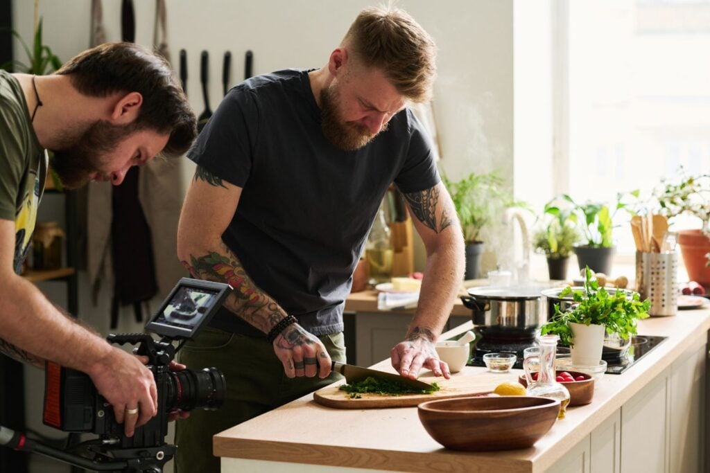 Two men in a kitchen - one cooking and one photographing