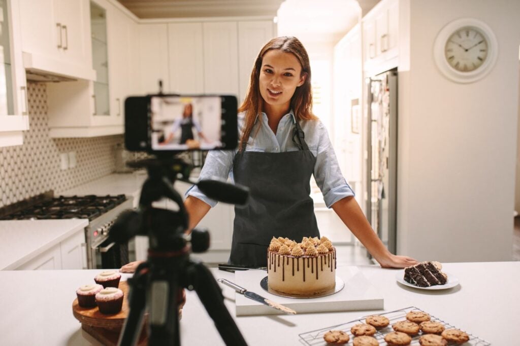 A woman talking to a camera with a cake and pastries on her kitchen counter